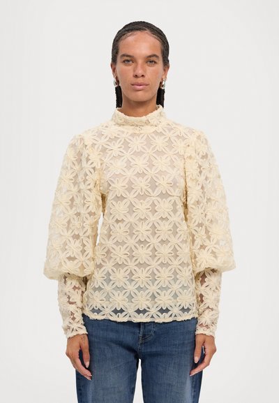 Kremfarget blondetopp med blomsterbroderi, høy hals og voluminøse ermer; kombinert med denimjeans. Strukturert, gjennomsiktig utseende.