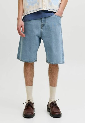 Osoba nosi svetlomodré riflové šortky, béžové ponožky a hnedé šnurovacie topánky, stojí s jednou rukou v vrecku na neutrálnom pozadí.