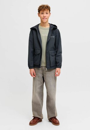 Tinejdžer s kovrčavom kosom u tamnoj kariranoj jakni, zelenoj majici, širokim sivim trapericama i smeđim kožnim cipelama, stoji i smiješi se.