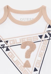 Bijela pamučna bodi sa bež ovratnikom. Ima istaknuti bež teksturirani upitnik okružen natpisom "GUESS" u crnoj i bež boji.