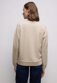 Sweat-shirt oversize beige avec encolure et poignets côtelés, texture lisse et coupe décontractée. Associé à un jean bleu foncé.