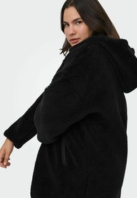 Melns, pūkains vilnas žakets ar kapuci, ar garām piedurknēm, brīvu piegriezumu un priekšējām kabatām. Mīksta tekstūra ar smalkām šuvju detaļām.