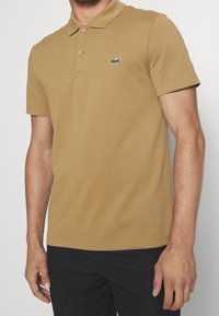 Polo tričko s krátkým rukávem v pískové bavlně s límečkem. Má dvě knoflíky, malé vyšívané logo na hrudi a texturovaný povrch.