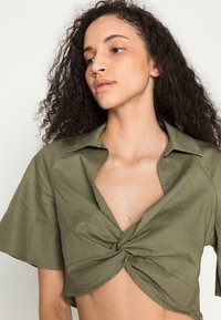 Camiseta curta em verde-oliva, feita de tecido texturizado, com um design de frente torcida e mangas curtas com colarinho.