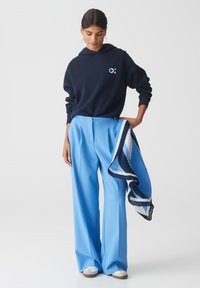 Marineblauwe hoodie met een logo, lichtblauwe wijde broek, roze en blauwe gestreepte sjaal, en lichte schoenen met een gestructureerde zool.