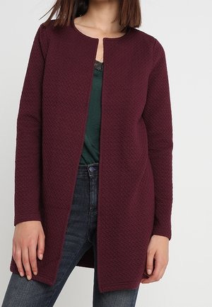 Femme portant un cardigan bordeaux texturé à manches longues et à ouverture frontale sur un top vert foncé et un jean bleu, sur un fond uni.