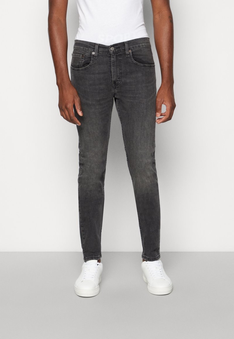 Jean en denim gris foncé coupe slim avec design à cinq poches, décoloration subtile et passants pour ceinture, associé à des baskets blanches.