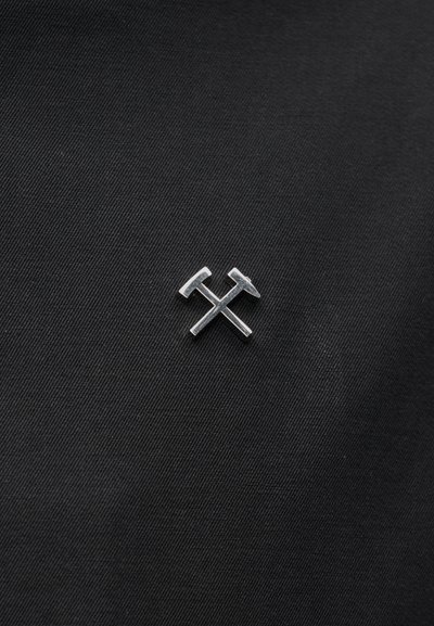 Silberne Nadel in Form von zwei gekreuzten Hämmern auf schwarzem, strukturiertem Stoffhintergrund.