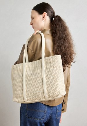 Kvinde med langt krøllet hår holder stor beige vævet skuldertaske, iført brun jakke og blå jeans på grå baggrund.