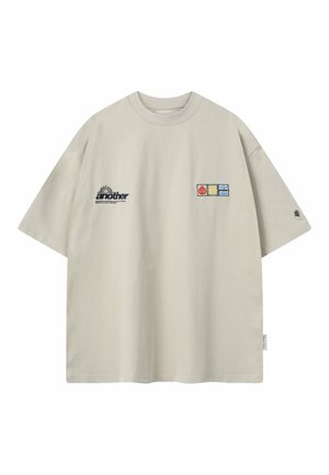 Camiseta beige de gran tamaño con mangas cortas, con el texto "another" y tres pequeños parches coloridos en el frente.