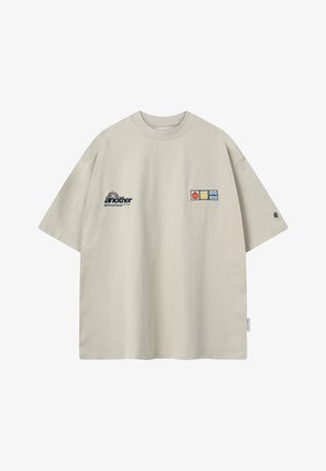 Camiseta beige de gran tamaño con mangas cortas, con el texto "another" y tres pequeños parches coloridos en el frente.