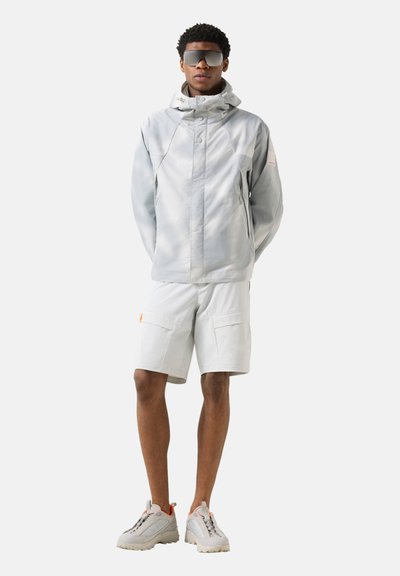 Młody mężczyzna ubrany w jasnoszarą kurtkę z kapturem, pasujące szorty typu cargo, masywne sneakersy i duże lustrzane okulary przeciwsłoneczne, stoi z rękami za plecami.