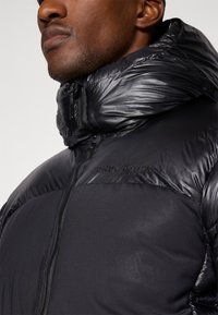Gros plan sur un homme portant une veste isolante noire de Peak Performance avec un col haut et une fermeture éclair à l'avant.
