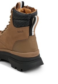 Bota de tobillo de ante marrón y nylon con cuello negro texturizado, ojales dorados y suela de goma negra. Cuenta con el logotipo de BOSS en el talón y el lateral.