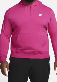 Růžová mikina Nike s přední kapsou, kapucí se stahovací šňůrkou a žebrovanými manžetami. Bílý logo na levé straně hrudníku. Materiál působí měkce a strukturovaně.