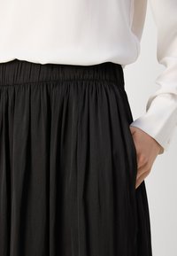 Svart plisserad kjol med elastisk midja, tillverkad av slätt tyg. Har sidofickor och är kombinerad med en vit blus med lång ärm.