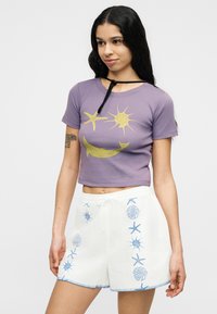 Νεαρή γυναίκα με μακριά σκούρα μαλλιά φοράει μωβ κοντό τοπ με κίτρινα θαλάσσια σχέδια και λευκά σορτς με μπλε κεντήματα εμπνευσμένα από τη θάλασσα.