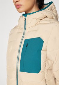 Beige dunjakke med en teal lomme, hette og glidelås i front. Har quiltet tekstur og en kontrastglidelås langs fronten.