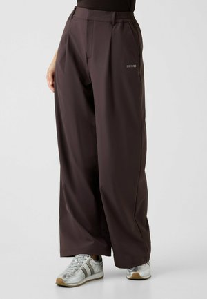 Donna che indossa pantaloni ampi di colore marrone scuro con il logo "GUESS" sulla coscia sinistra e sneakers argento con lacci bianchi.