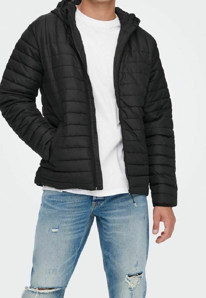 Homme portant une doudoune noire sur une chemise blanche et un jean bleu clair déchiré, les mains dans les poches de la veste.