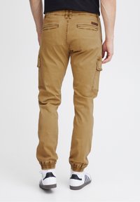 Kaki cargobyxor med elastiska fållar, med fickor på låren och en brun läderlogotyp på baksidan. Slät bomullstyg.