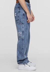 Blue denim jeans with a relaxed fit, faded wash, and embroidered branding on the pocket. Worn with white sneakers.