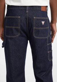 Vista trasera de una persona vistiendo pantalones de mezclilla azul oscuro con grandes bolsillos y un parche con el logo de Guess en el bolsillo trasero derecho y la cintura.