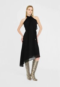 Schwarzes asymmetrisches Kleid aus leichtem Stoff mit Rüschenakzenten und hohem Ausschnitt. Kombiniert mit metallisch schimmernden kniehohen Stiefeln im Schlangenledermuster.