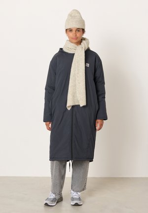 Tumši zila polsterēta jaka ar pilna garuma rāvējslēdzēju, kopā ar gaiši pelēku adītu cepuri un šalli, un gaišām džinsām. Izskatu papildina sporta apavi.