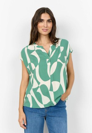 Kobieta z długimi ciemnymi włosami, ubrana w zieloną i białą bluzkę z krótkim rękawem o abstrakcyjnym wzorze oraz niebieskie dżinsy, stoi z jedną ręką w kieszeni.