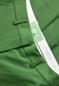Groene stoffen broek met een witte tailleband en een groene label met "JXX." Heeft platte zakken en een gladde textuur.