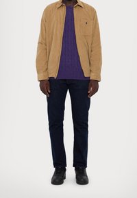 Persona che indossa una giacca beige con cerniera, maglione lavorato a trecce viola, jeans blu scuro e scarpe da ginnastica nere, in piedi davanti a uno sfondo bianco.