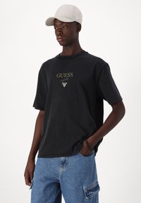 Svart bomull T-skjorte med løs passform, med gull "GUESS" logo og trekantete emblem. Båret med blå denim cargo-bukser og en lys caps.
