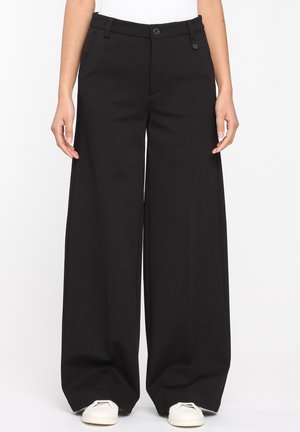 Nainen pukeutuneena korkeavyötäröisiin, leveälahkeisiin mustiin housuihin, joissa on etupuolella napit ja vetoketju, yhdistettynä valkoisiin lenkkareihin, seisomassa yksiväristä taustaa vasten.