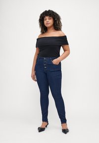 Top negro off-the-shoulder combinado con jeans ajustados azul oscuro que cuentan con un cierre frontal de botones y bolsillos laterales, usado con planos negros de punta.