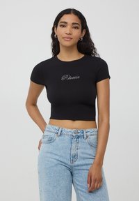 Czarny krótki t-shirt z miękkiego materiału z krótkimi rękawami, z białym tekstem z rhinestone "Rêverie" na środku. Noszony z jasnoniebieskimi dżinsami.