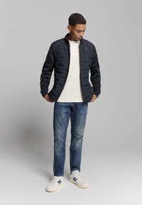Navy gebokt jasje met hoge kraag, gecombineerd met een beige t-shirt, blauwe jeans en witte sneakers met marineblauwe accenten. Casual ontwerp.