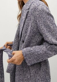 Blazer tessuto di colore viola e bianco con un motivo testurizzato, colletto a risvolto e un dettaglio con cravatta in vita. Presenta due tasche frontali e chiusura con bottoni.