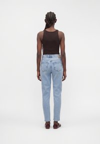 Persona con cabello largo trenzado que lleva una camiseta marrón sin mangas, vaqueros azul claro y zapatos burdeos, de pie frente a un fondo blanco.