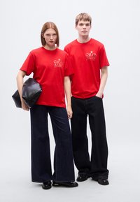 Κόκκινα βαμβακερά t-shirts με το λογότυπο Calvin Klein, συνδυασμένα με σκούρα τζιν παντελόνια φαρδιάς γραμμής· ένα μοντέλο κρατάει ένα μαύρο δερμάτινο clutch.