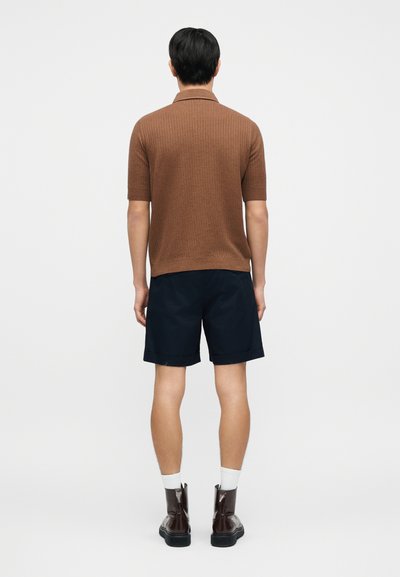 Polo marron côtelé à manches courtes, associé à un short bleu marine, avec des bottines marron et des chaussettes blanches, vu de dos.