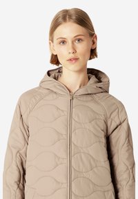 Chaqueta acolchada con capucha de color marrón claro, con un patrón de olas, que presenta un cierre frontal de cremallera y bolsillos laterales. Textura suave y diseño minimalista.