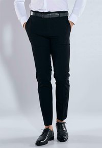 Schwarze Anzughose mit schmaler Passform, glatter Textur, Gürtelschlaufen und schlichtem Design. Kombiniert mit polierten schwarzen Schuhen.