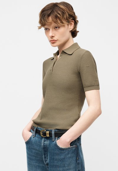 Άτομο με κοντά κυματιστά μαλλιά που φοράει κοντομάνικη πόλο μπλούζα ελιάς και μπλε τζιν με μαύρη ζώνη, τα χέρια στις τσέπες, ουδέτερο φόντο.