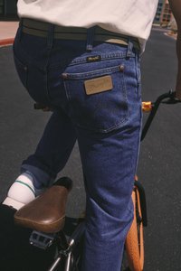 Inimene, kes kannab siniseid Wrangler-jeanse ja valgeid tossusid, sõidab oranžratastega jalgrattaga asfalditeel.