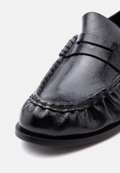 Mocassin noir en cuir brillant avec bout mocassin cousu et détail de bride à la barrette sur fond blanc.