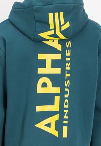 Soodne teal kapuutsiga, mille seljal on suur kollane trükk, mis ütleb "ALPHA INDUSTRIES" julge, kaasaegse fontiga. Pehme kanga tekstuur.