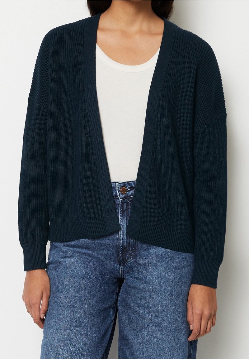 Kvinna klädd i en mörk marinblå stickad kofta över en vit T-shirt och högmidjade blå jeans med avslappnade händer längs sidorna.