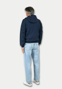 Giacca a vento blu navy con texture liscia; jeans azzurri; scarpe bianche con accenti neri; vista da dietro, che mostra i dettagli dell'indumento.