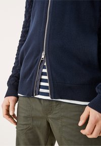 Granatowy sweter na zamek z pikowanym detalem na ramionach, ściągaczami na mankietach i dole. Pod spodem częściowo widoczna jest pasiasta koszula.